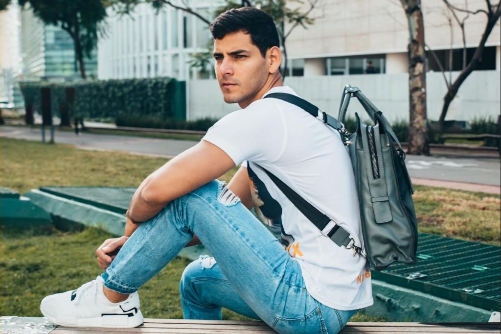 Young man sitting on a bench in a modern urban park, wearing a casual outfit featuring a white graphic t-shirt, ripped light blue jeans, and white sneakers. He carries a stylish gray leather backpack with sleek straps, exuding a contemporary and relaxed vibe.