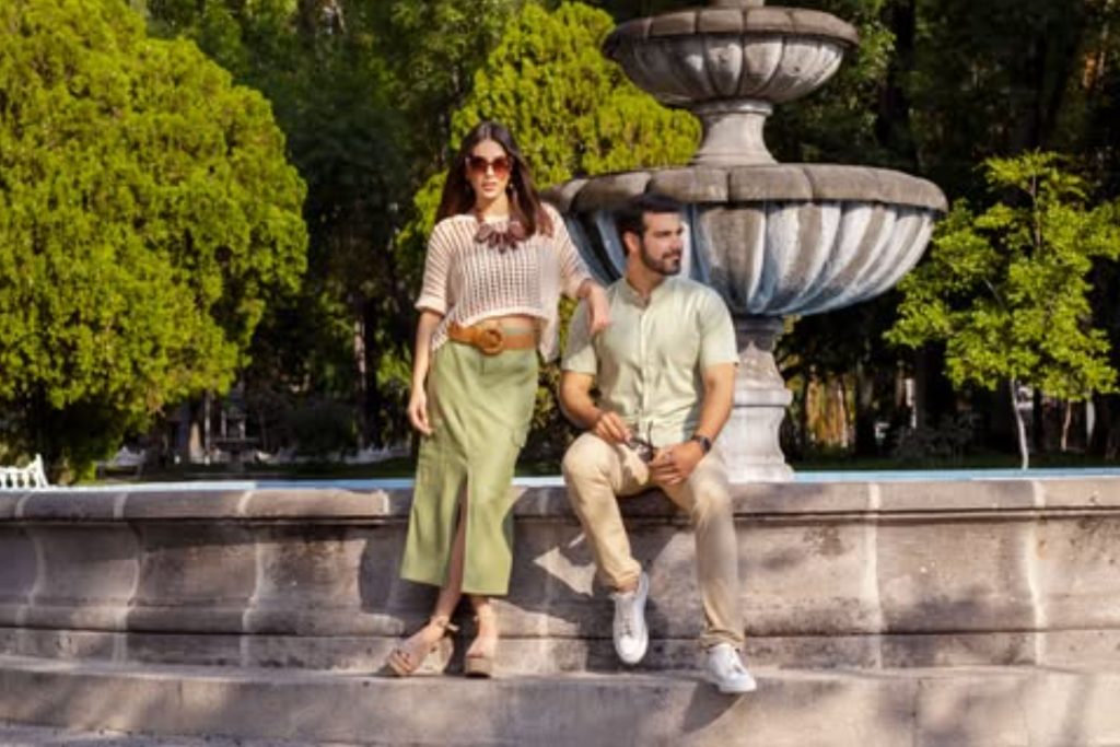 Stylish man and woman posing in front of a large stone fountain in a lush park. The woman wears a chic knit top, a high-waisted green skirt, and wedge sandals, accessorized with sunglasses and a statement belt. The man is dressed in a light green shirt, beige pants, and white sneakers, holding a pair of sunglasses, creating a relaxed yet fashionable look.