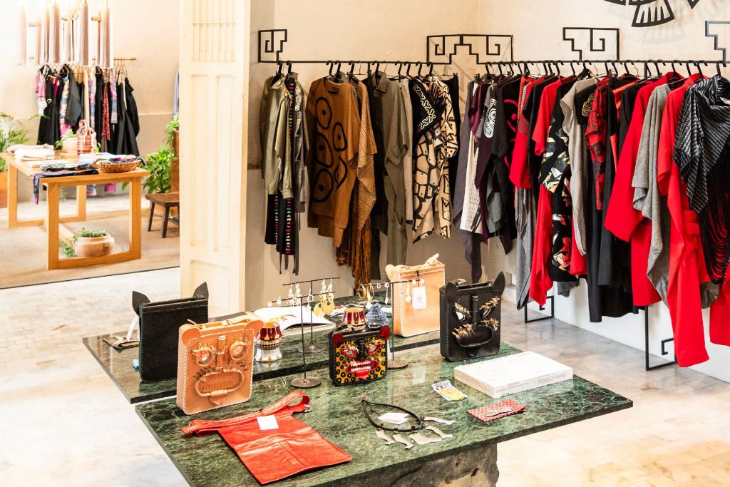 Interior of a boutique featuring traditional and modern Mexican clothing. Racks with ponchos, jackets, and dresses in earthy and bold colors are displayed, along with a table showcasing handmade accessories and decorative items. The store design incorporates cultural motifs.