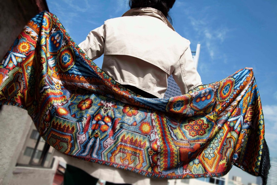 A vibrant silk scarf with intricate Mexican designs featuring floral and geometric patterns held up by a person wearing a beige coat. The colorful scarf contrasts against a clear blue sky in the background.