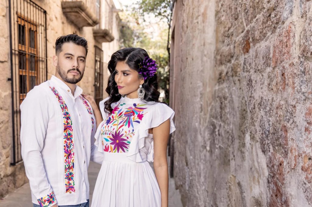 Man and woman wearing traditional Mexican embroidered clothing. The man is dressed in a white guayabera-style shirt with colorful floral embroidery, while the woman wears a white dress adorned with vibrant floral patterns, complemented by statement earrings and a purple flower in her hair. They stand in a rustic stone alleyway with soft natural lighting.